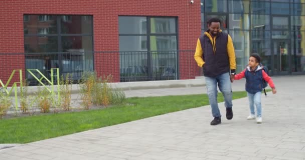 Sevgi dolu bir baba ve sırt çantalı, atlayan ve okuldan yürüyerek gelen tatlı bir oğul. Neşeli Afrikalı Amerikalı baba ve genç çocuk öğrenci eğleniyor ve oynuyorlar. Aile ilişkileri hedefi. Eğitim kavramı — Stok video
