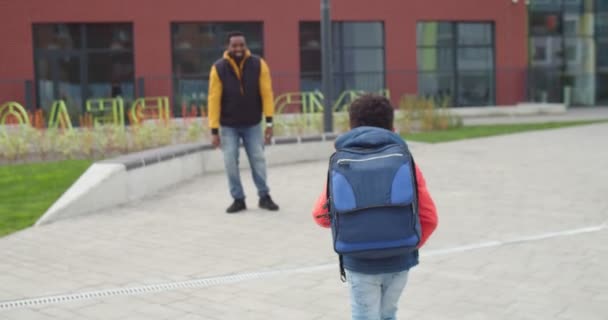 Parte trasera de un niño afroamericano con mochila que va desde la escuela hasta un padre feliz y cariñoso. Primer plano de padre amoroso guapo abrazando pequeño colegial lindo al aire libre. Concepto de alumno feliz — Vídeos de Stock