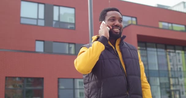 Retrato de un hombre afroamericano alegre parado en la calle y sonriendo mientras llama y habla por teléfono celular. Hombre guapo feliz cerca de la escuela charlando en el teléfono inteligente de buen humor. Concepto urbano — Vídeos de Stock