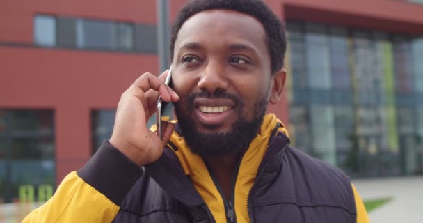 Veselý Afroameričan stojí na ulici a usmívá se, zatímco volá a mluví na mobilu. Šťastný pohledný muž blízko školy mluví na smartphonu v dobré náladě. Zavřít koncept portrétu — Stock video