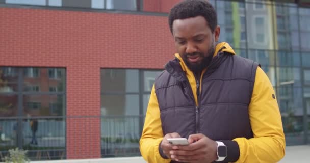 Porträt eines gut aussehenden afroamerikanischen Mannes, der allein auf der Straße geht und Probleme mit der Internetverbindung hat, während er SMS auf dem Handy schreibt. Männchen tippen auf Smartphone und versuchen, Signal im Freien zu finden — Stockvideo