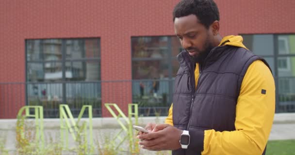 Портрет радісного афроамериканця, який йде вулицею і має проблеми з Інтернетом під час смс на мобільному телефоні. Чоловіки стукають по смартфону і намагаються знайти сигнал, посміхаючись — стокове відео