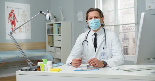 Egy jóképű fehér férfi orvos portréja orvosi maszkban és fehér kabátban, sztetoszkóppal a szekrényben ülve a kórházban, a koronavírus alatt, és a kamerába nézve. Egészségügyi koncepció — Stock videók