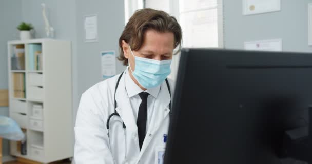 Médecin masculin professionnel caucasien occupé en masque médical et manteau blanc assis au bureau dans l'armoire de la clinique, tapant et tapant sur l'ordinateur sur le lieu de travail. Ferme là. Concept hospitalier — Video