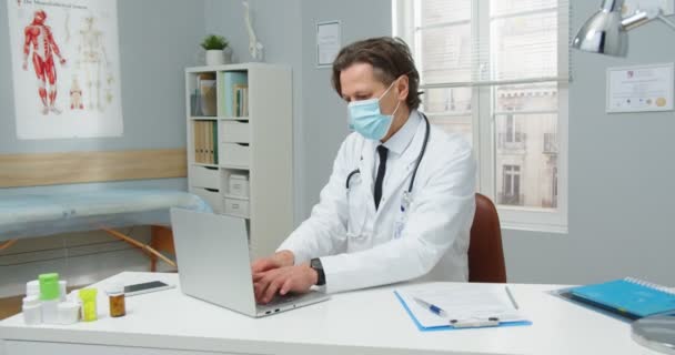 Drukke serieuze blanke professionele man arts met medisch masker zitten aan tafel in het ziekenhuis kast, werken aan de computer, tikken en typen op laptop op het werk. Covid-19 pandemie, portretconcept — Stockvideo