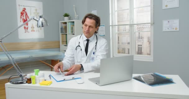 忙碌而严肃的身穿医疗白衣的白人职业医生坐在诊所橱柜的办公桌前，一边用智能手机说话，一边在工作场所用笔记本电脑打字。医院概念 — 图库视频影像
