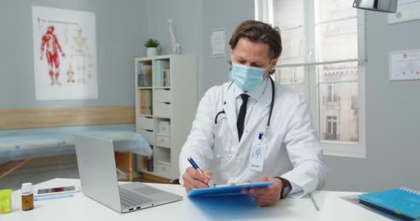 Portrét zaneprázdněného bělošského profesionálního lékaře v lékařské masce, který sedí za stolem v nemocniční skříni, pracuje, píše na notebooku a píše léčbu v dokumentech. Covid-19 pandemický koncept — Stock video