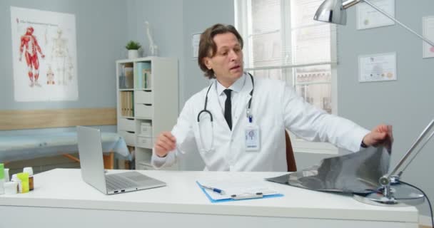 Porträt eines Arztes mittleren Alters in medizinischem Mantel, der am Tisch im Krankenhauszimmer sitzt, Röntgenbilder betrachtet und Ergebnisse in Dokumente schreibt. Konzept der Coronavirus-Pandemie — Stockvideo