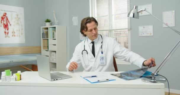 Porträt eines umtriebigen kaukasischen Arztes mittleren Alters in medizinischem Mantel, der im Krankenhauszimmer am Laptop sitzt und die Röntgenergebnisse betrachtet. Coronavirus-Pandemie. Behandlungskonzept — Stockvideo