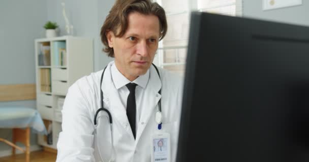 코카서스의 한 중년의 의사가 병원 캐비닛에 앉아 컴퓨터로 타이핑하고, 일하고 모니터를 보고 있습니다. 치료 개념. 의료 전문가 — 비디오
