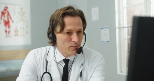 Primer plano de guapo de mediana edad feliz hombre caucásico médico especialista en atención médica sentado en la clínica hablando en videollamada en el ordenador con auriculares que tienen consulta en línea con el paciente. Concepto de salud — Vídeos de Stock