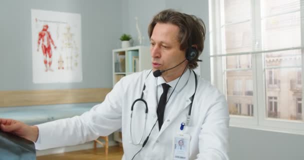 Porträt eines gutaussehenden kaukasischen Arztes mittleren Alters, der im Krankenhauszimmer sitzt, Video-Chat im Headset mit Online-Konsultation, die dem Patienten die Diagnose erklärt — Stockvideo