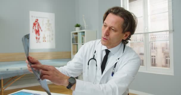 Portrait of middle-aged Caucasian male healthcare specialist general practitioner sitting in hospital room video chatting having online consultation on internet explaining X-ray scan to patient. — Stock Video