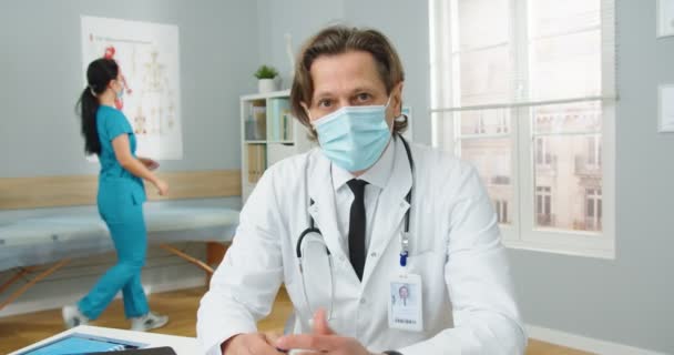 Vista en primera persona. Retrato del guapo médico caucásico en máscara médica sentado en el gabinete de la clínica y explicando el tratamiento, hablando en video llamada consulta en línea pandemia Covid-19 — Vídeos de Stock