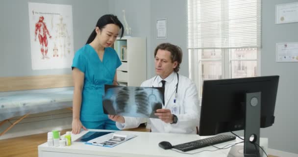 白种人中年有经验的男医生坐在诊所的橱柜里，与亚洲女性助手谈论病人的诊断、 X光扫描。治疗结果。肺病工作概念 — 图库视频影像