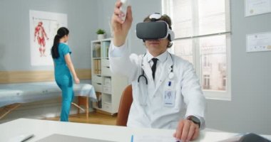 Uzman beyaz erkek hekimin portresi. Klinikte oturmuş sanal gerçeklik cihazıyla el ele dolaşırken Asyalı kadın hemşire arka plandaki belgeleri alıyor.