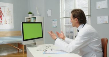 Kafkasyalı erkek sağlık uzmanının klinikteki masasında oturması, internet kamerasından yeşil ekrana bakması. Krom anahtarlı monitör. çevrimiçi danışma