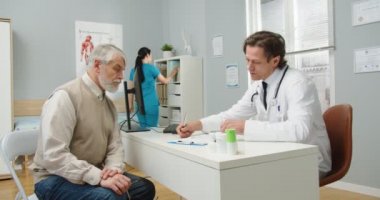 Klinik kabinesindeki beyaz erkek doktorun yan görüşü yaşlı bir hastayla hastanede tedavi hakkında konuşurken ve reçete yazarken. Asyalı kadın hemşire arka planda çalışıyor.