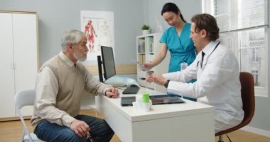 Yan görüş. Kafkasyalı orta yaşlı profesyonel erkek doktor hastanede oturuyor ve Asyalı kadın hemşire için imza atarken kıdemli erkek hastayla hastalık tedavisi hakkında konuşuyor. Klinik kavramı