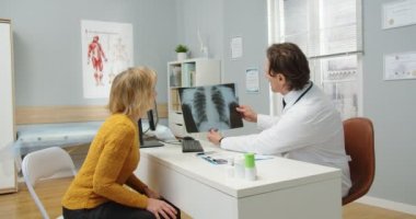 Klinikteki yakışıklı erkek doktorun yan görüntüsü masada oturan, yaşlı hasta kadınla konuşan ve hastanede röntgen taramasını gösteren. Asyalı kadın hemşire arka planda çalışıyor.
