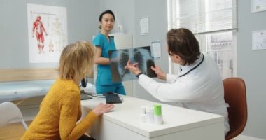 Yan görüş. Klinikte oturan orta yaşlı beyaz erkek doktorun portresi, Asya 'lı kadın hemşire ona analiz sonuçlarını verirken, yaşlı kadın hasta ile konuşuyor.