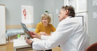 Kafkasyalı profesyonel doktor masasında oturmuş, yaşlı kadın hastayla röntgen taramasını açıklarken ve yorumlarken konuşuyor. Beyin cerrahı konsepti. Sağlık Hizmetleri