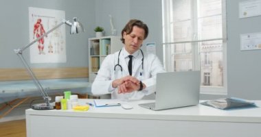 Ciddi orta yaşlı beyaz erkek sağlık uzmanının portresi hastane odasında oturuyor, çalışıyor ve bilgi yazıyor. Dizüstü bilgisayara bakıyor ve hasta tıbbı konseptini bekliyor.