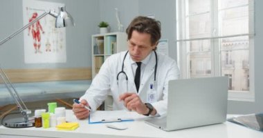 Orta yaşlı, beyaz, yakışıklı bir erkek doktorun portresi hastane ofisinde masasında oturuyor, saatine bakarken belgelere bakıyor ve yazıyor. Tedavi kavramı reçeteleniyor