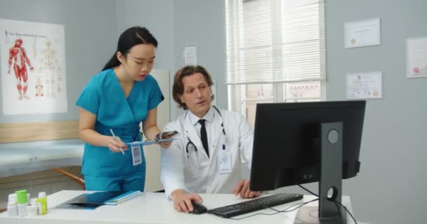 Портрет кавказького фахівця з охорони здоров'я, який сидить у кабінеті лікаря в медичному центрі і друкує на комп'ютері, розмовляє з азіатською жінкою-асистентом про діагноз пацієнта, результати лікування. — стокове відео