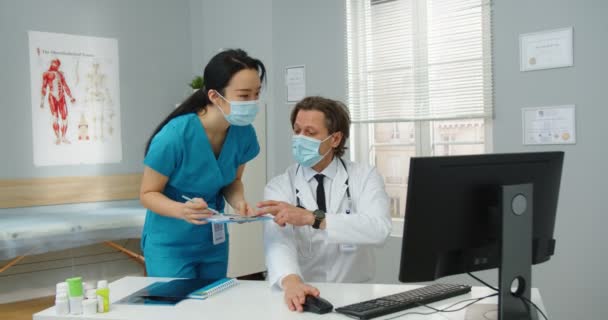 Retrato de hombre caucásico especialista en atención médica profesional en máscara sentado en la habitación en el centro médico y tocando en la computadora, discutiendo con la mujer asiática asistente de enfermería resultados de tratamiento de pacientes — Vídeos de Stock