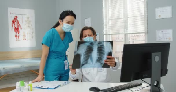 Specjaliści od opieki zdrowotnej w maskach medycznych omawiający skan rentgenowski, wyniki analiz. Lekarz wyjaśniający leczenie młodej azjatyckiej pielęgniarce. Koncepcja koronawirusa. zakażenie płuc, wir 19 — Wideo stockowe