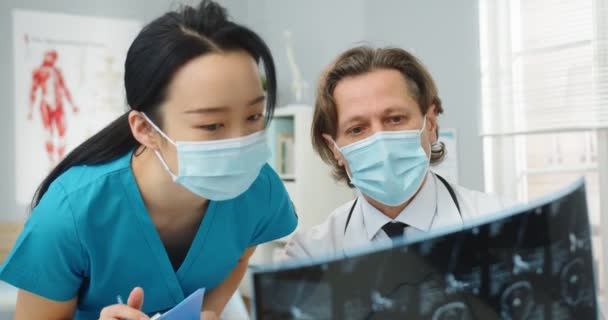 Primer plano de los neurólogos profesionales de la salud multiétnicos en máscaras médicas discutir la exploración de rayos X, resultados de análisis. Médico varón comentando diagnóstico a enfermera asiática joven. Concepto de medicina — Vídeo de stock