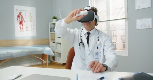 Portret profesjonalnego lekarza kaukaskiego, specjalisty w dziedzinie opieki zdrowotnej w okularach VR siedzącego w gabinecie klinicznym i poruszającego się ręka w rękę z wykorzystaniem innowacyjnej technologii wirtualnej rzeczywistości w medycynie — Wideo stockowe