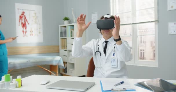 Портрет профессионального врача-кавказца в очках VR, сидящего в больничном кабинете и делающего жесты, используя инновации виртуальной реальности, в то время как азиатская медсестра ходит на заднем плане — стоковое видео