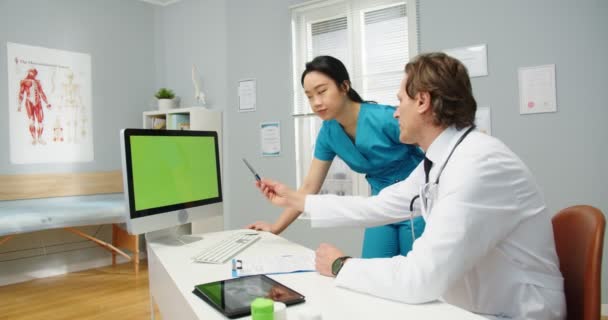 Seitenansicht des kaukasischen Mannes Gesundheitsspezialist Arzt sitzt im Kabinett im medizinischen Zentrum und schaut auf Computer grünen Bildschirm, im Gespräch mit asiatischen Assistentin über Patientendiagnose, Chroma-Schlüssel — Stockvideo