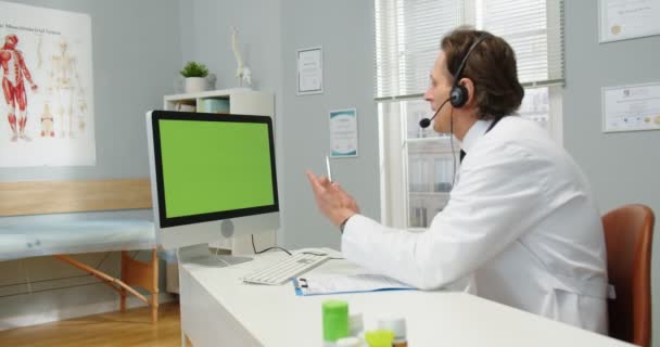 Oldalnézetből. Kaukázusi férfi orvos portréja fülhallgatóban, amint az asztalnál ül a kórházi szekrényben, videokamerázik videóhívással, miközben webkamera figyeli a zöld képernyőt. Monitor krómkulccsal — Stock videók