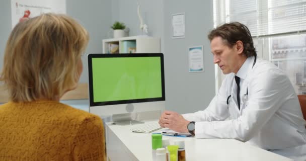 Vista posterior de la paciente mujer escuchando al médico caucásico de mediana edad sentado en el escritorio en el hospital y explicando los resultados del diagnóstico mirando la pantalla de la computadora con pantalla verde, concepto de retrato — Vídeos de Stock