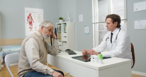 白种人老年男性患者与男性医生预约时坐在医院橱柜中的侧视图。亚洲女护士提供医生证明。专家向病人解释治疗情况 — 图库视频影像