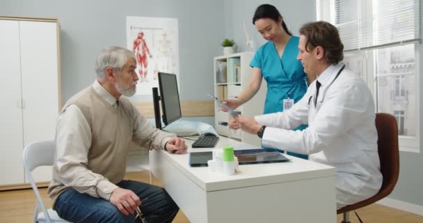 Vista lateral. Médico masculino profesional caucásico de mediana edad sentado en el hospital y hablando con un paciente adulto mayor sobre el tratamiento de la enfermedad mientras firma papeles para la enfermera asiática. Concepto clínico — Vídeos de Stock