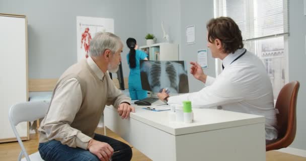 Vista lateral de un especialista en salud masculino caucásico de mediana edad en el hospital en el escritorio, previa consulta con un paciente anciano que explica y muestra una radiografía. Enfermedad pulmonar. Concepto Covid-19 — Vídeos de Stock