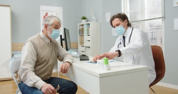 Boční pohled na bělocha profesionální lékař v lékařské masce na klinice sedí za stolem, mluví se starším mužem pacient předepisuje léčbu a dávat pilulky v nemocnici. covid-19 koncept — Stock video