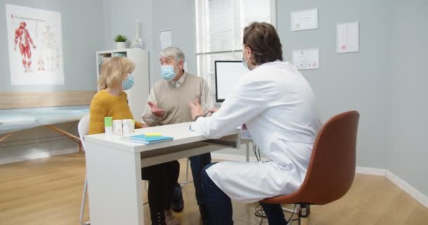 Coppia di anziani caucasici seduti in ospedale su consultazione medica in chat con il medico che sta prescrivendo e dando loro pillole e medicine. L'infermiera lavora sullo sfondo. Concetto di pandemia di coronavirus — Video Stock