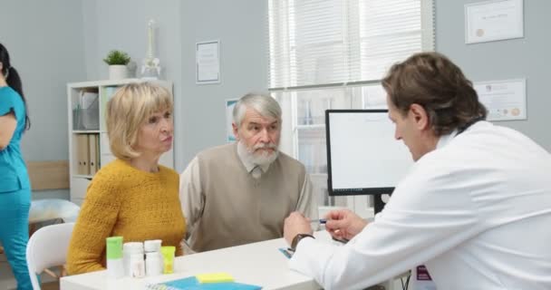 Portrét starého bělošského manželského páru sedícího na klinice na lékařské konzultaci u lékaře a hovořícího s mužským specialistou. Doktor dává pilulky starším pacientům. Koncept zdravotní péče — Stock video