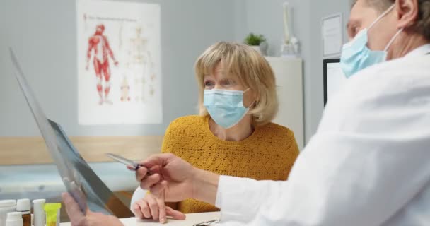 Vista posterior del médico caucásico en máscara médica discutiendo y explicando la exploración por rayos X a una paciente mayor de sexo femenino en consulta en el consultorio del hospital. problema de salud, pandemia de Coronavirus, Primer plano — Vídeos de Stock