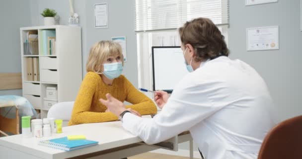 코카서스에서는 하얀 코트와 의료용 마스크를 착용 한 중년 남성 의사가 병원 캐비닛에 있는 나이든 여성 환자와 건강 문제에 대해 논의하고 있다. 코로나 바이러스 대유행 , Covid-19 컨셉트 — 비디오