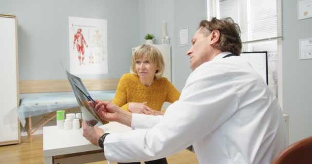 Rear of Caucasian man professionele arts in het medisch centrum zit aan het bureau, spreken met oude vrouw patiënt op overleg uitleggen en becommentariëren X Ray scan. Neurochirurg concept. Gezondheidszorg — Stockvideo