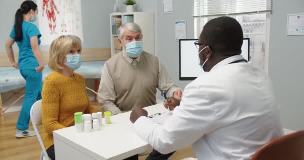 Ritratto di coppia anziana caucasica uomo e donna in maschere mediche seduti in clinica su consultazione medica con il medico afroamericano maschio. Posteriore del medico uomo che parla con i pazienti — Video Stock