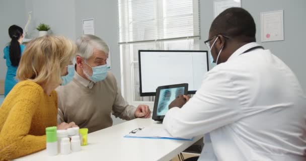 Primer plano del médico afroamericano en mascarilla pinchando en la tableta que muestra la exploración por rayos X a pacientes viejos. Pareja de ancianos caucásicos sentados en el hospital en consulta médica. Concepto de tratamiento — Vídeos de Stock