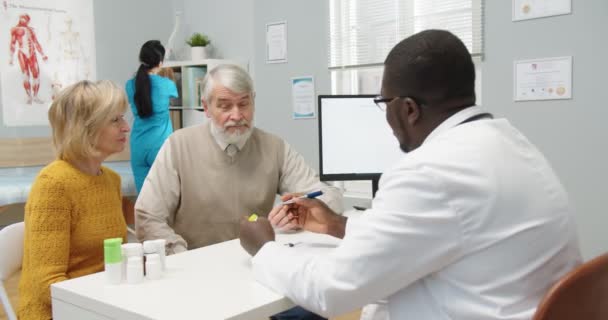 Klinikte tıbbi danışmanlık yapan ve Afrikalı Amerikalı erkek doktor uzmanla konuşan yaşlı, beyaz, evli bir çiftin portresi. Nörologun arkası ilaç ve hap veriyor. — Stok video