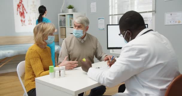 Achterzijde van Afro-Amerikaanse arts in masker voorschrijven behandeling en het geven van pillen aan oude man. Oudere blanke getrouwde echtpaar zit in het ziekenhuis kamer op doktersafspraak. Coronavirus concept — Stockvideo